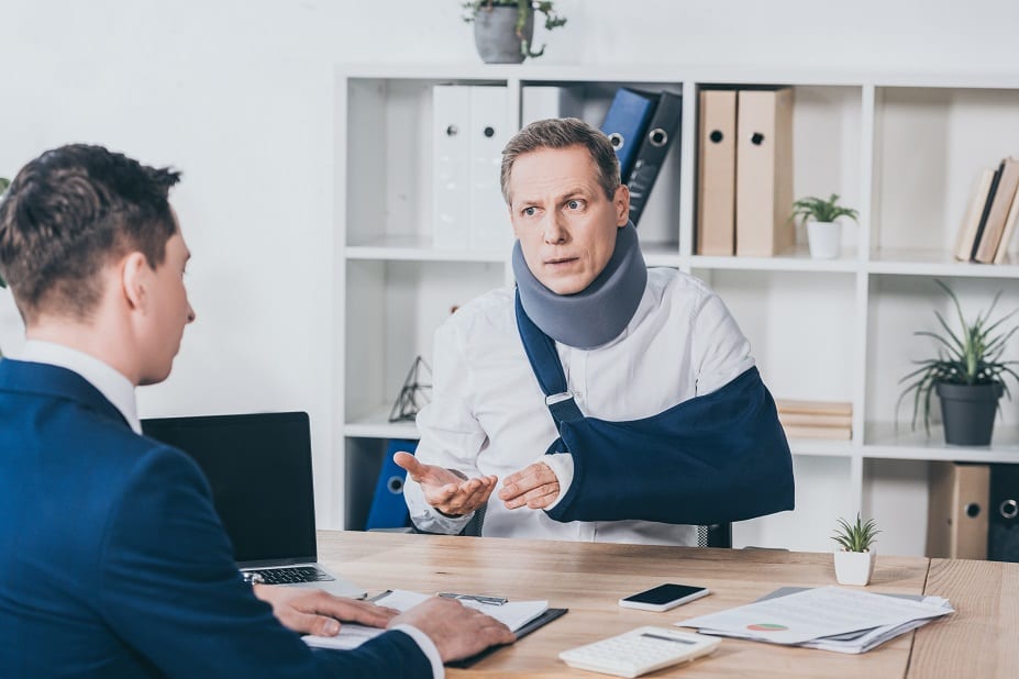 middle aged worker in neck brace with broken arm sitting at table and talking to businessman in blue jacket in office, compensation concept