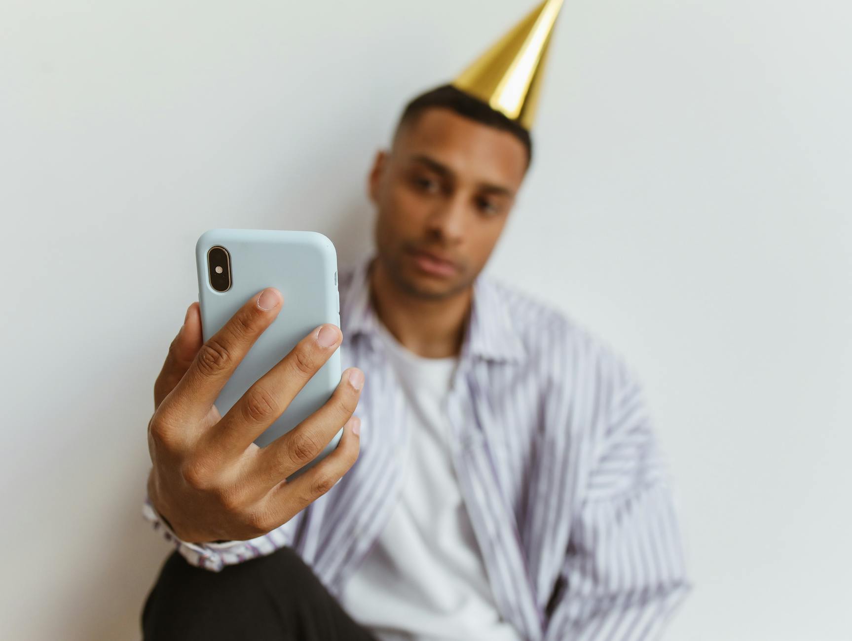 A man wearing a party hat takes a selfie with his smartphone indoors against a plain white background.