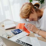 A woman overwhelmed with finances, surrounded by bills, calculator, and cash at home.