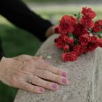 Delicate hands resting on headstone with red carnations, symbolizing remembrance.