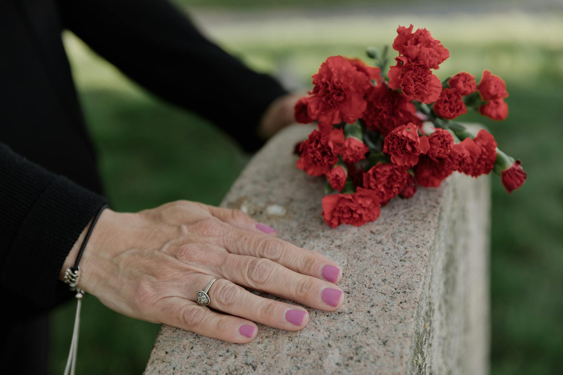 Delicate hands resting on headstone with red carnations, symbolizing remembrance.