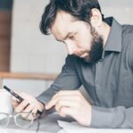 A bearded man intensely stares at his smartphone, deep in thought, indoors.