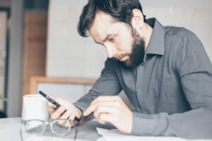 A bearded man intensely stares at his smartphone, deep in thought, indoors.