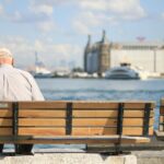An elderly man sits alone on a bench by the waterfront, enjoying a peaceful day.