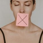 Young woman in black lingerie with closed eyes showing pink sticker with black cross against violence and abuse on lips on white background in light studio