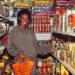 Happy African American lady in casual outfit with shopping basket in hand choosing food from products in small supermarket among colorful boxes on shelves and smiling with mouth opened