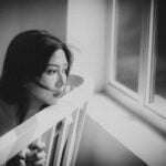 Black and white depressed ethnic female sitting on chair and looking out window at home