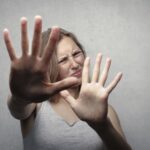 Portrait of a fearful woman in a gray tank top with hands pushed forward against a gray background.
