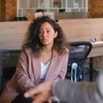 A woman sitting at an office desk listening calmly to a colleague who is gesturing toward her