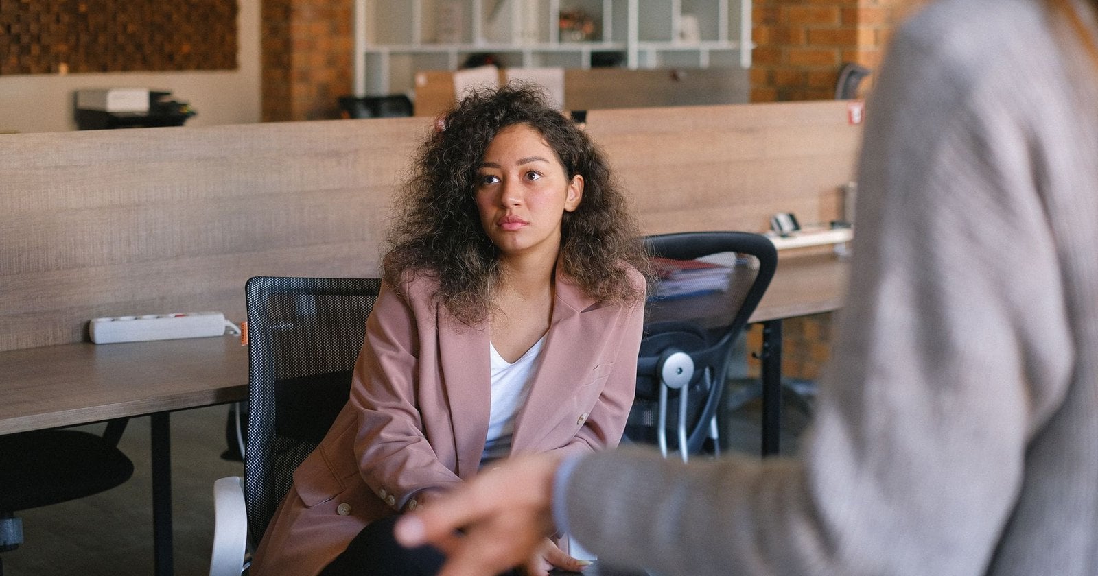 A woman sitting at an office desk listening calmly to a colleague who is gesturing toward her